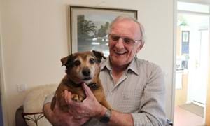An elderly gentleman smiles while he holds his small pet dog, who is looking at the camera.