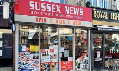 A small shopfront with glass windows and doors, and a red sign with Sussex News in big white bold text