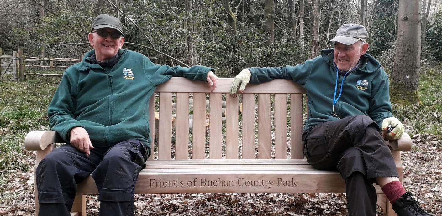 nojs Volunteers - Two volunteers sat on a wooden bench with the words 'Friends of Buchan Country Park' engraved.