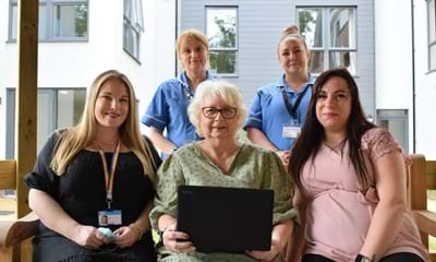 Two members of care staff at an Extra Care facility stand behind two members of staff and a resident who are sat on a bench with one of the donated laptop computers.