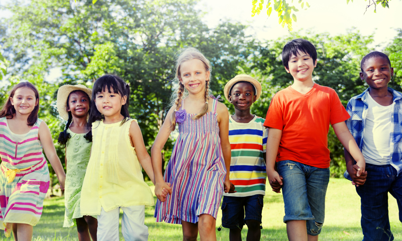Four young girls and three young boys in a line, holding hands and smiling as they walk towards the camera.