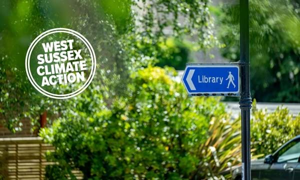 Library sign on pole with greenery and bushes and West Sussex Climate Action go in white
