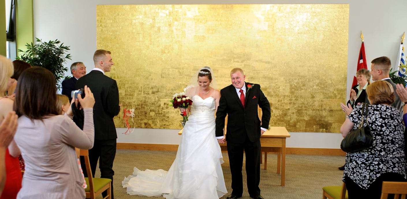 nojs Smiling bride and groom standing at the front of the ceremony room in front of a gold painting