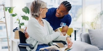 Young male caregiver in scrubs and elderly woman in wheelchair enthusiastically speaking together.