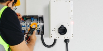 Close-up of a male electrician in a hard hat and high-vis vest performing tests with a clamp meter during home EV charger installation. 