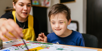 Young female teacher painting  at a school desk with a schoolboy with special educational needs. 