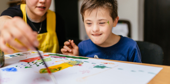 Young female teacher painting  at a school desk with a schoolboy with special educational needs. 