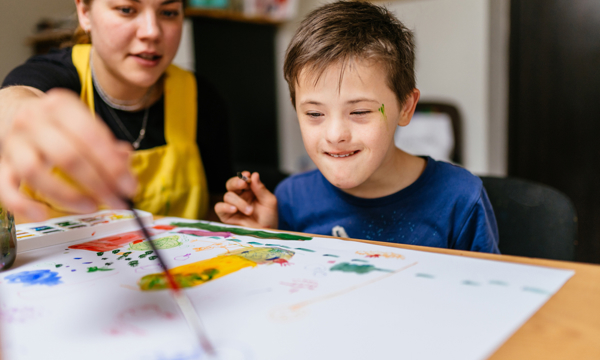 Young female teacher painting  at a school desk with a schoolboy with special educational needs. 