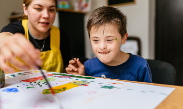 Young female teacher painting  at a school desk with a schoolboy with special educational needs.