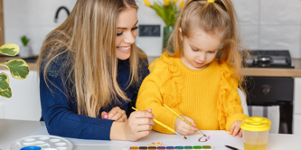 Young woman and small girl in yellow jumper painting pictures in a home setting.