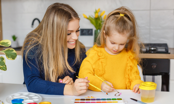 Young woman and small girl in yellow jumper painting pictures in a home setting.
