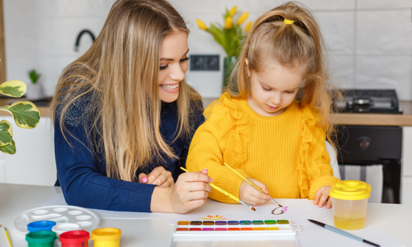 Young woman and small girl in yellow jumper painting pictures in a home setting.