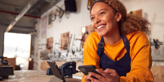 Happy young female employee in a workshop environment, wearing an apron and using a digital tablet while holding mobile phone. 