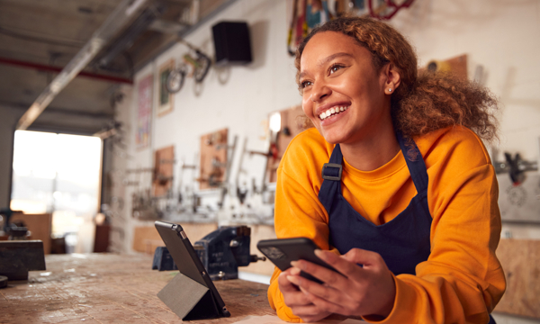 Happy young female employee in a workshop environment, wearing an apron and using a digital tablet while holding mobile phone. 