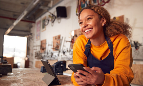 Happy young female employee in a workshop environment, wearing an apron and using a digital tablet while holding mobile phone.