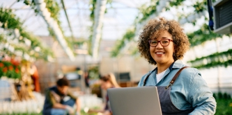 Happy female garden plant nursery worker using laptop and looking at camera.