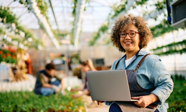 Happy female garden plant nursery worker using laptop and looking at camera.
