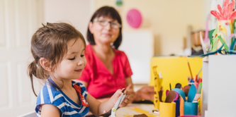 Middle-aged early years teacher sitting with young girl supervising crafts, as the girl uses a pair of child’s scissors.  
