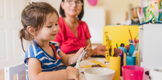 Middle-aged early years teacher sitting with young girl supervising crafts, as the girl uses a pair of child’s scissors.  