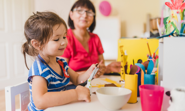 Middle-aged early years teacher sitting with young girl supervising crafts, as the girl uses a pair of child’s scissors.  
