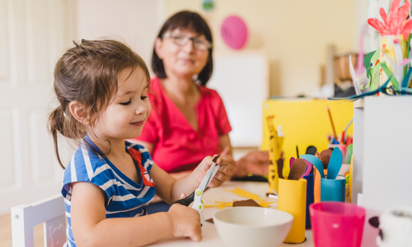 Middle-aged early years teacher sitting with young girl supervising crafts, as the girl uses a pair of child’s scissors.
