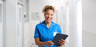 Female nurse in uniform standing in hospital corridor with clipboard smiling 