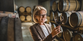 Mature female winemaker standing in winery holding clipboard and glass of wine