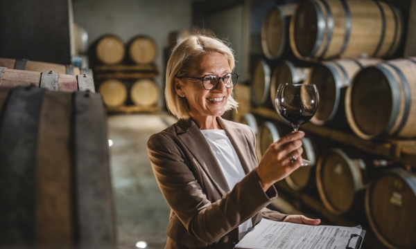Mature female winemaker standing in winery holding clipboard and glass of wine