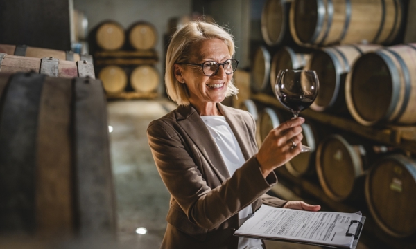 Mature female winemaker standing in winery holding clipboard and glass of wine