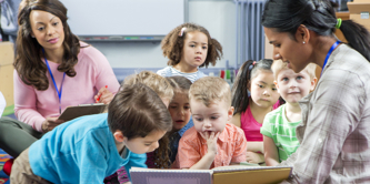 Young female pre-school teacher reads a story. The class is sitting on the floor. A female supervisor takes notes. 