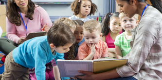 Young female pre-school teacher reads a story. The class is sitting on the floor. A female supervisor takes notes. 