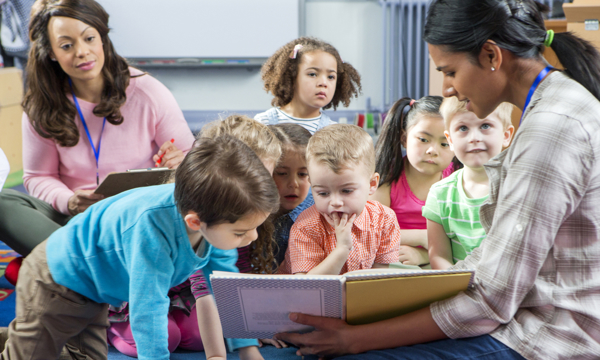 Young female pre-school teacher reads a story. The class is sitting on the floor. A female supervisor takes notes. 