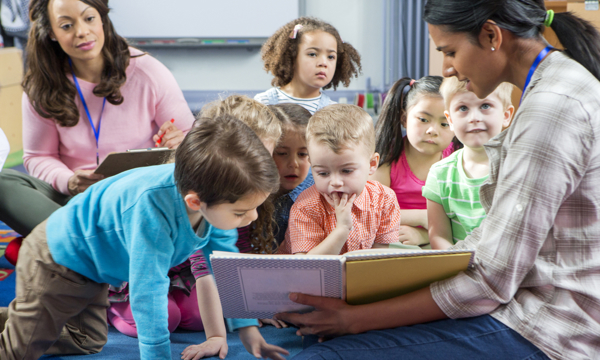 Young female pre-school teacher reads a story. The class is sitting on the floor. A female supervisor takes notes. 
